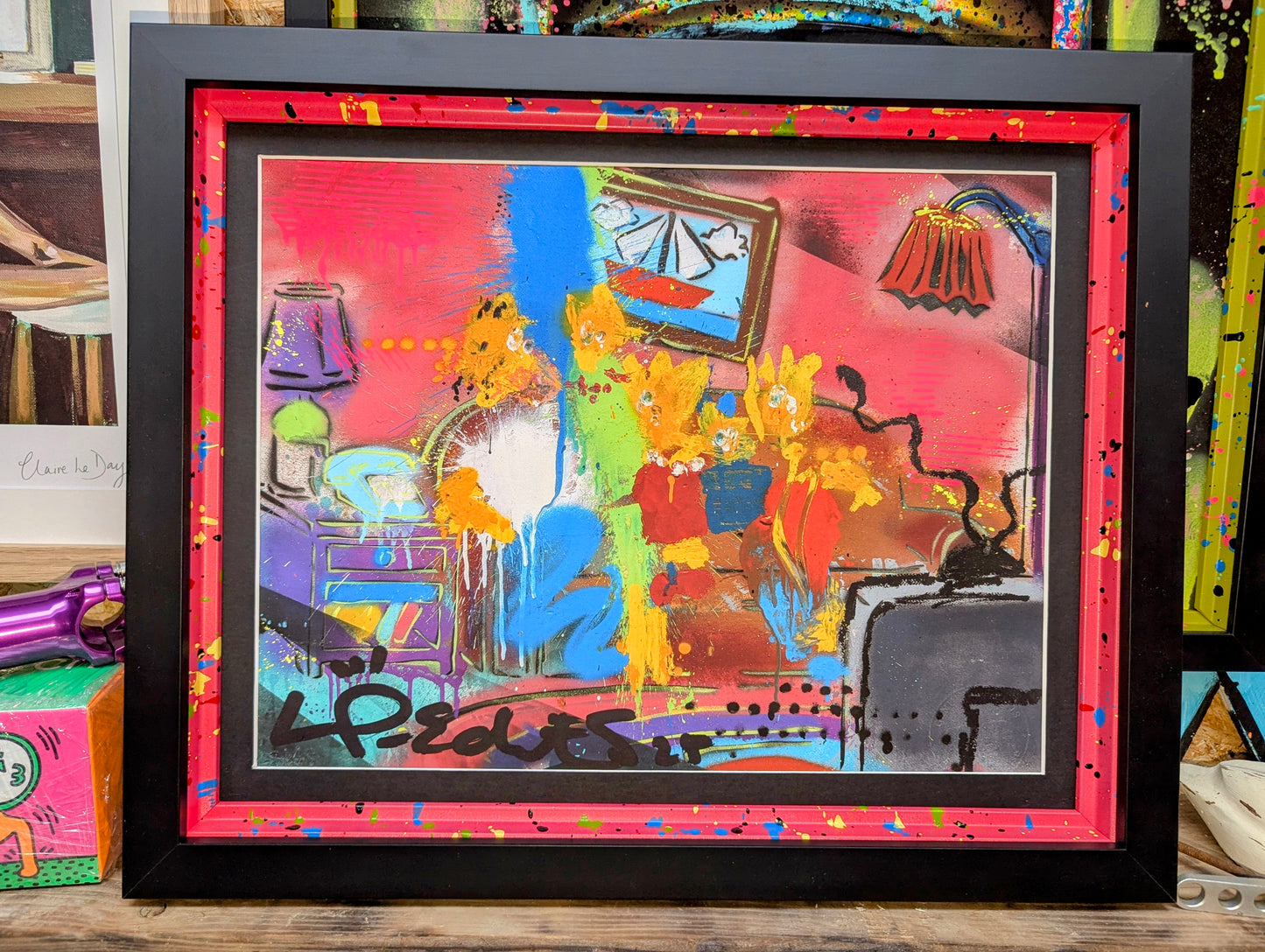 Lp edits artwork framed in a smooth black frame, painted internal frame painted red with blue yellow blue splatters. Artwork is Lp edits chaotic style of The Simpsons, frame is on wooden shelf surrounded by art
