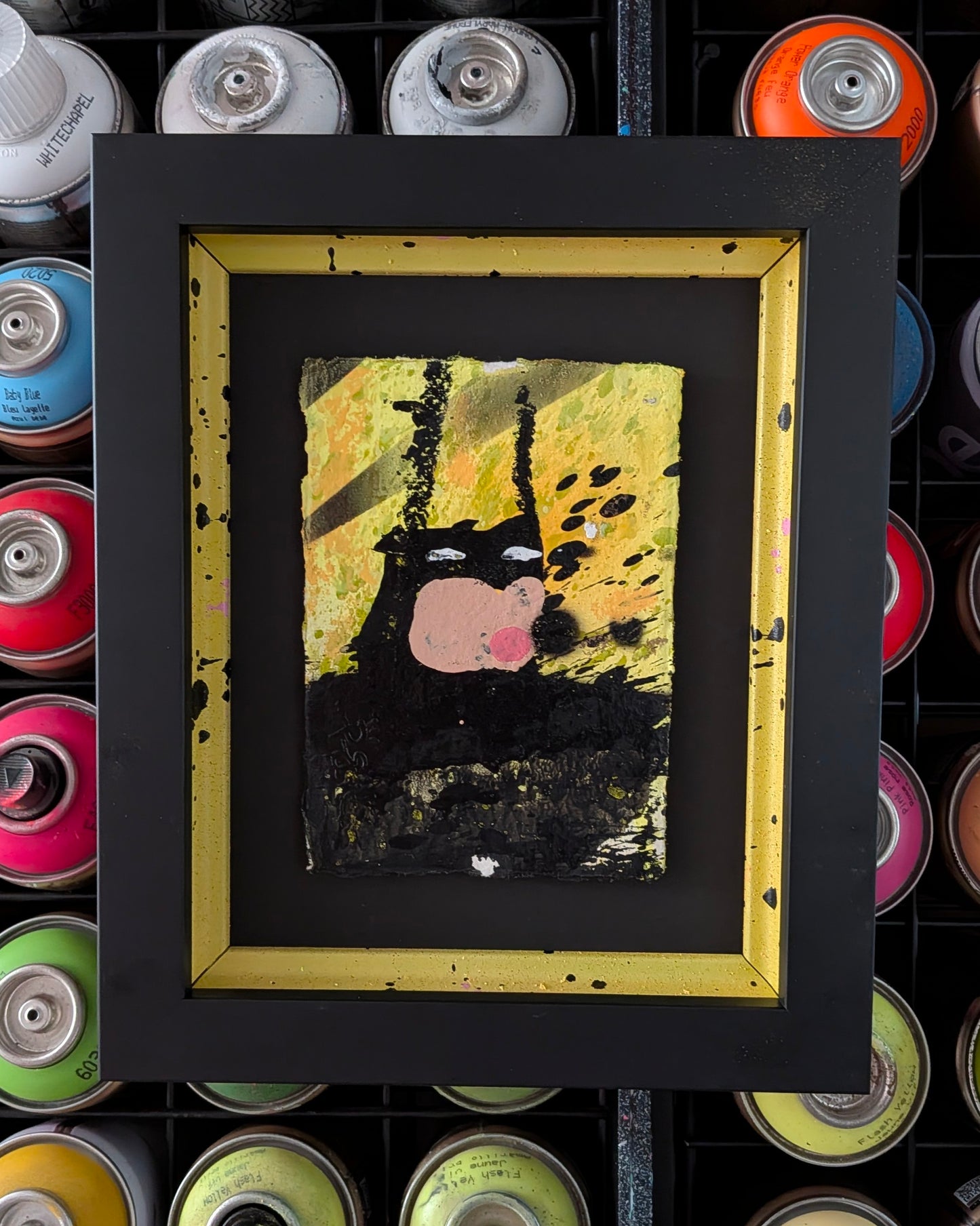 A framed abstract art print featuring a stylized black and pink face on a textured yellow and black background, set in a black frame with a gold inner border, displayed against a background of colorful spray paint cans.