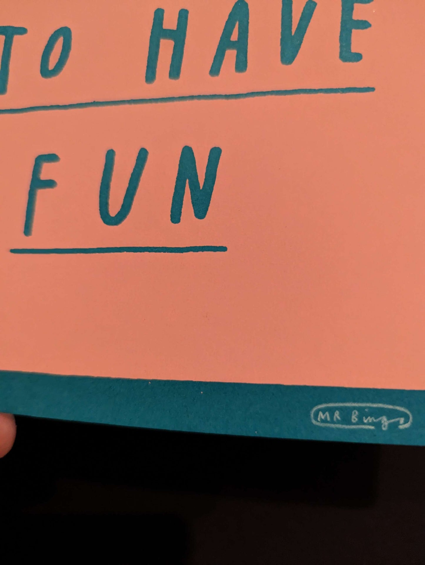 MR BINGO 'DON'T FORGET TO HAVE FUN' Ltd Ed Print, text is hand written style on a salmon pink tombstone against a teal background. card is being held in bottom left corner against a black background, this is a close up of Mr Bingos Signature