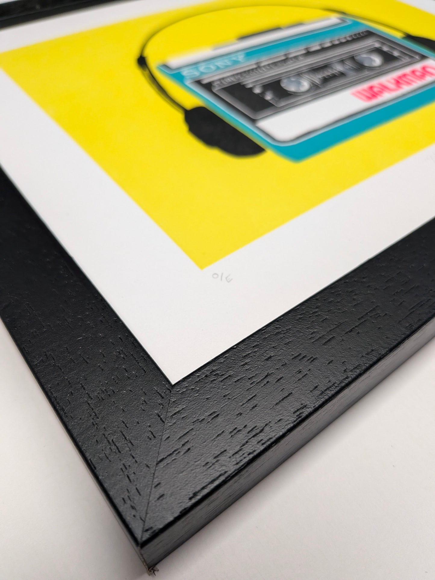 A framed art print featuring a retro cassette tape illustration with a bright yellow background, white matting, and a textured black frame.