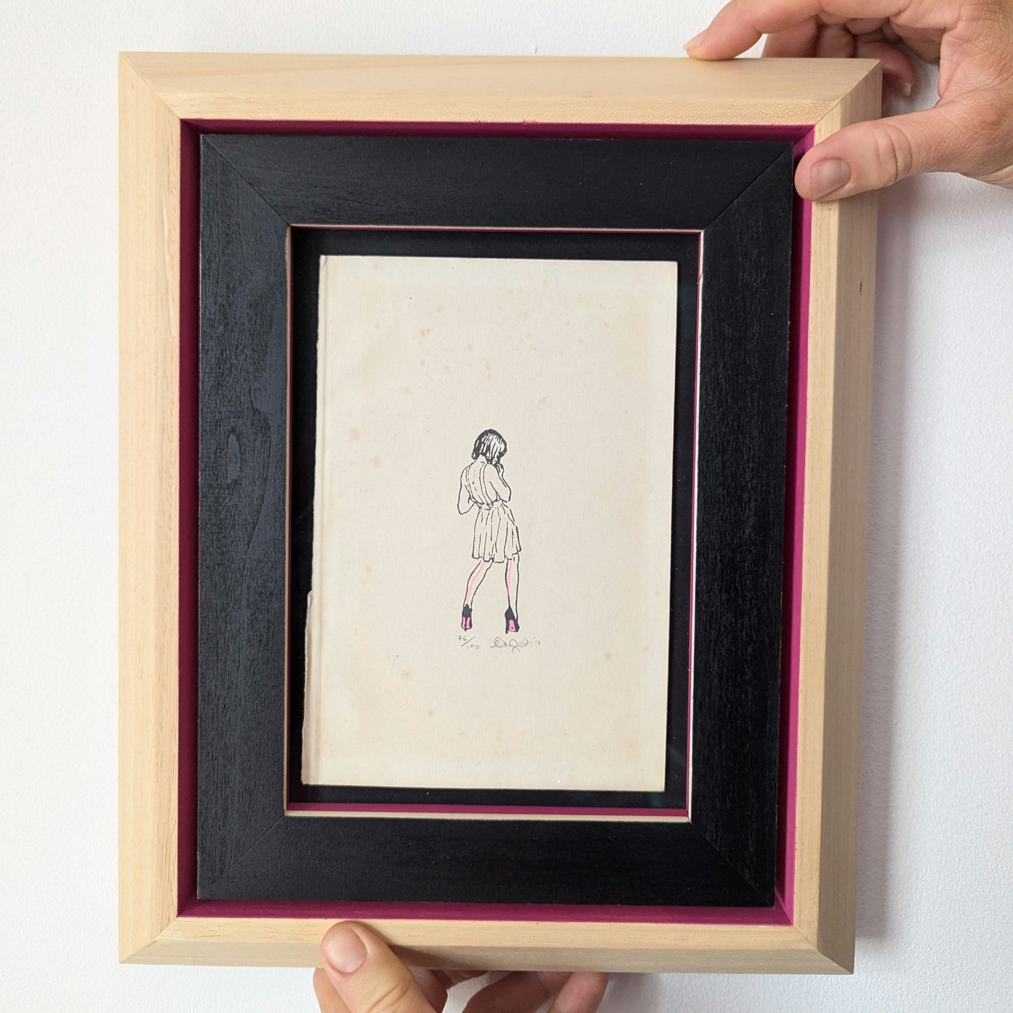 CEAL WARNANTS High Fashion artwork being held, artwork is in a wood frame with black and pink colouring, artwork is drawn on old book paper, the drawing is of a girl wearing a dress, her back is to us and she has pink details to her dress.