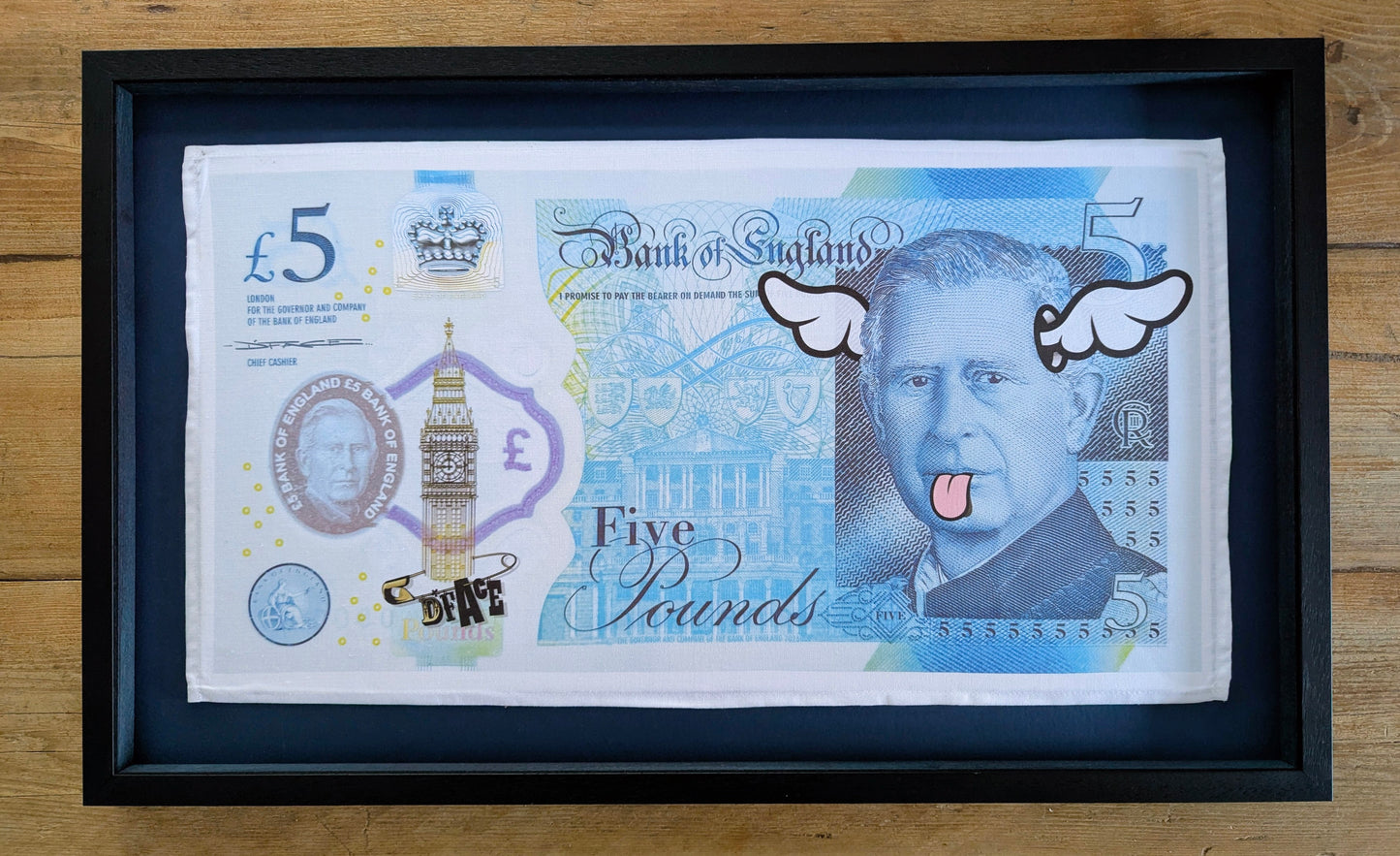 Framed Bank of England five-pound note tea towel with cartoonish face and wings on wood background
