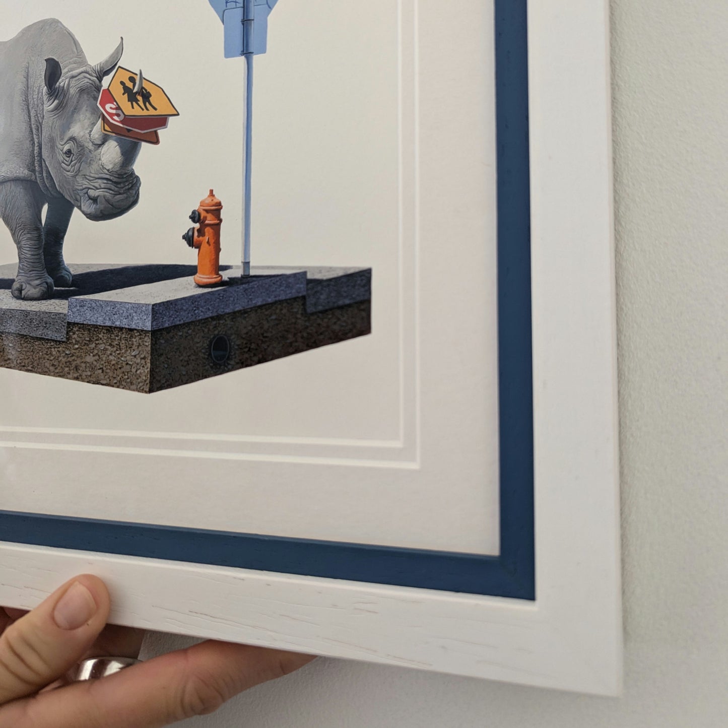 Framed artwork of a rhino with a street sign and fire hydrant on a platform, hand holding bottom of the frame