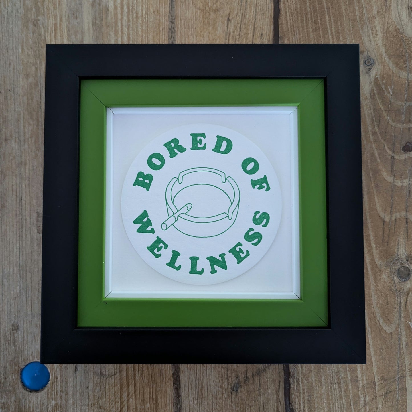 bored of wellness beer mat, green text with image of ashtray and lit cigarette, beer mat is float mounted on a double frame, internal frame is green and black out frame,