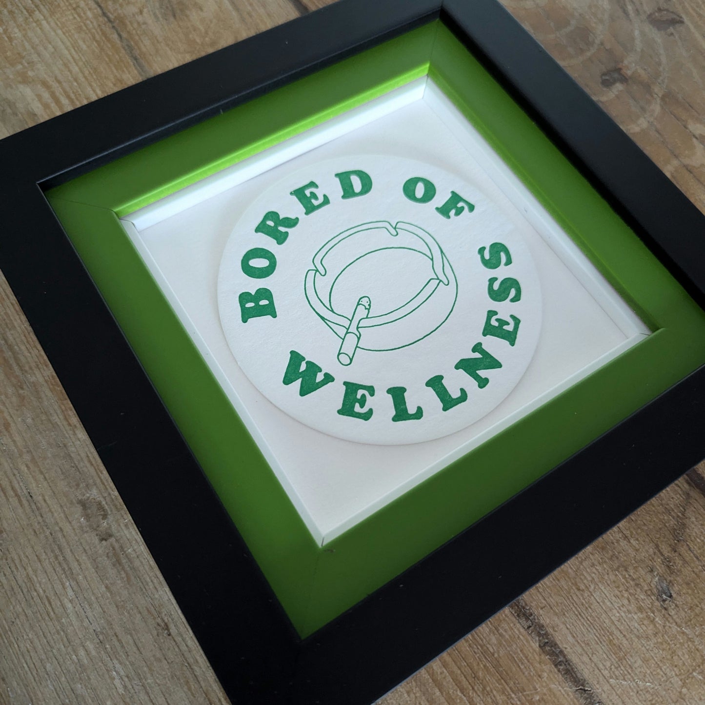 bored of wellness beer mat, green text with image of ashtray and lit cigarette, beer mat is float mounted on a double frame, internal frame is green and black out frame, angle photo.