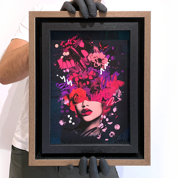 olly howe artwork, collage style womans face lips and nose, eyesare replaced by flowers and squiggles. framed in black frame, black frame is in brown float frame with black shadow gap. picture frame is being held by person in jeans and white shirt with black gloves.