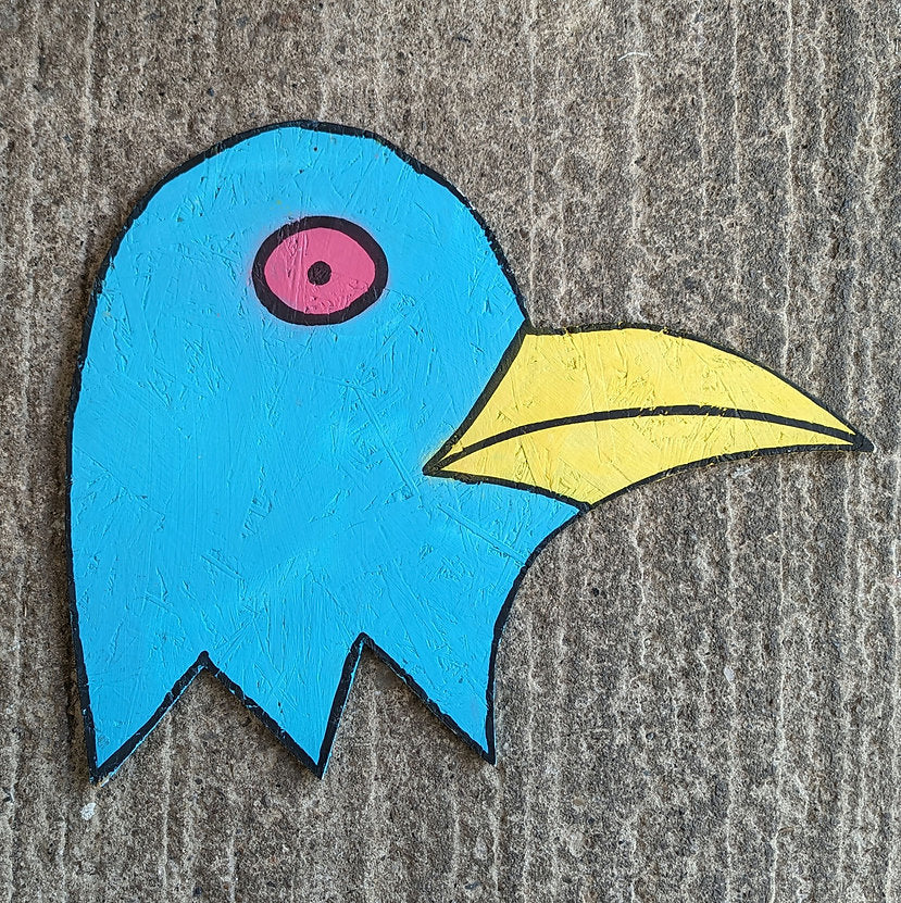 Steve McCracken blue bird head cut out of wood, cartoon shaped bird head painted in blue with pink eye and yellow beak. background is on concrete