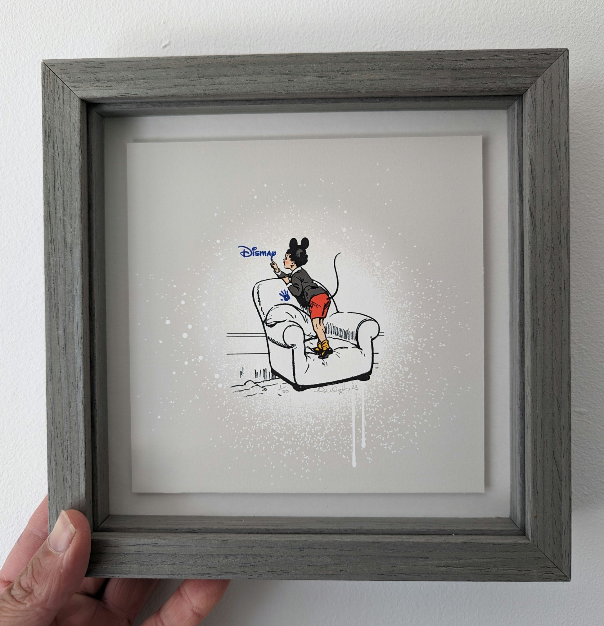 CHILD WITH MICKEY MOUSE EARS AND A TAIL STOOD ON A COMFY LOOKING ARMCHAIR, BLUE HAND PRINT ON CHAIR AND WRITING DISMAY IN DISNEY STYLE WRITING, IN A GREY PICTURE FRAME