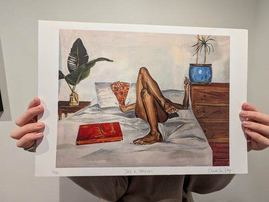 Art print of a person lying on a bed  eating a pizza slice next to pizza box with a plant and nightstand. being held by a person against white background.