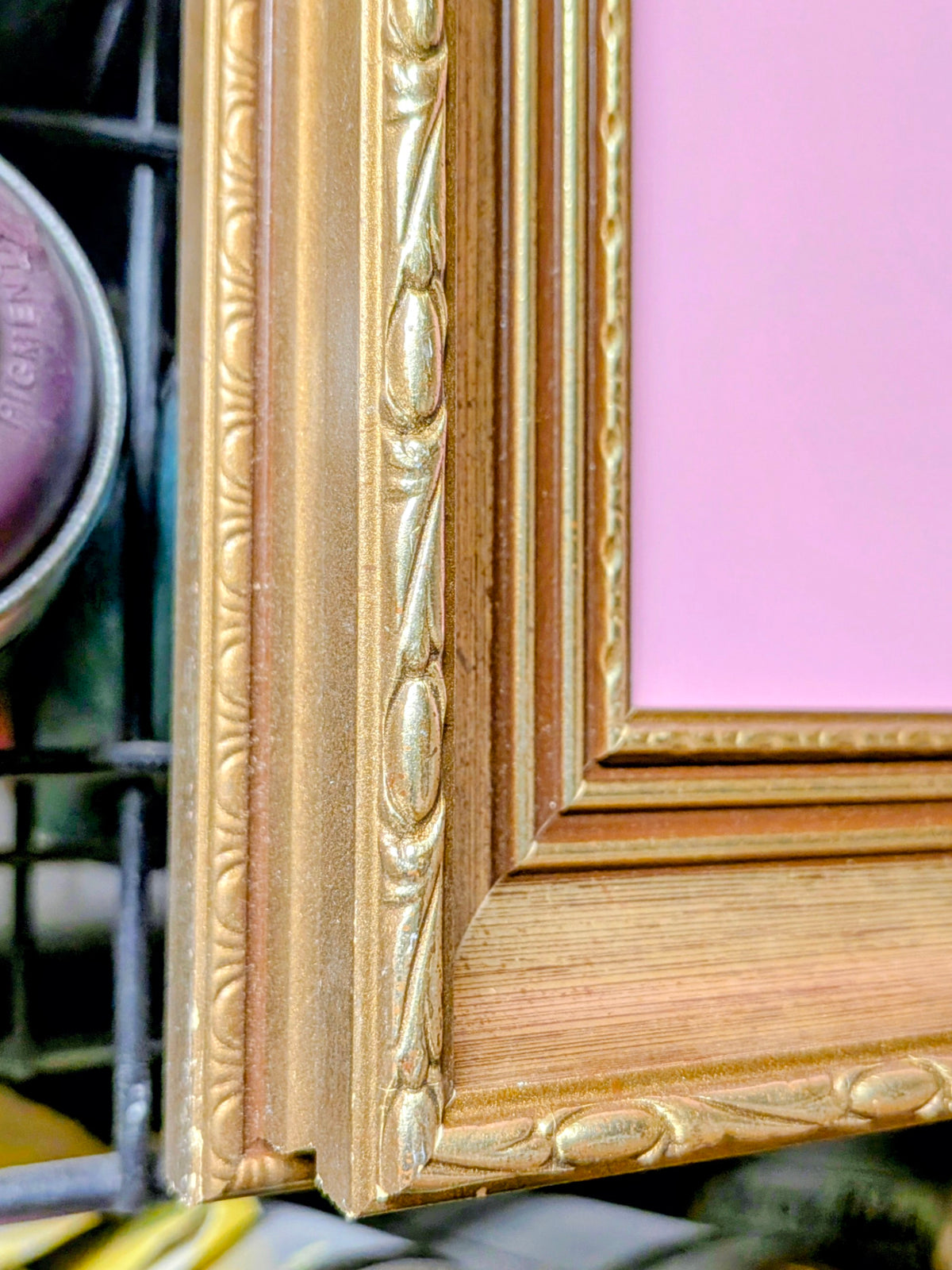 Close-up of an ornate gold picture frame corner with a pink insert or backing visible, photographed against a dark shelving background.