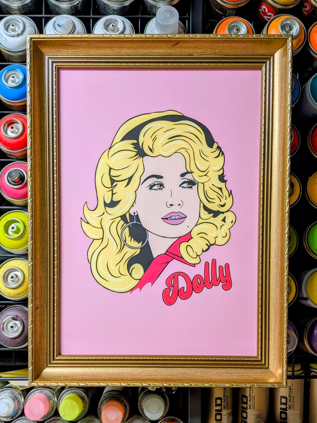 A framed pop art portrait print of Dolly Parton, featuring her signature blonde hair, red jacket, and pink background, enclosed in a gold frame and displayed against a backdrop of colorful spray paint cans.