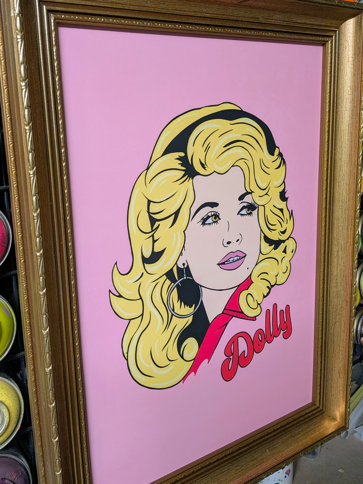 A framed pop art print featuring a stylized portrait of Dolly Parton with yellow hair, a pink background, and the name 'Dolly' in red script, shown in a gold frame.