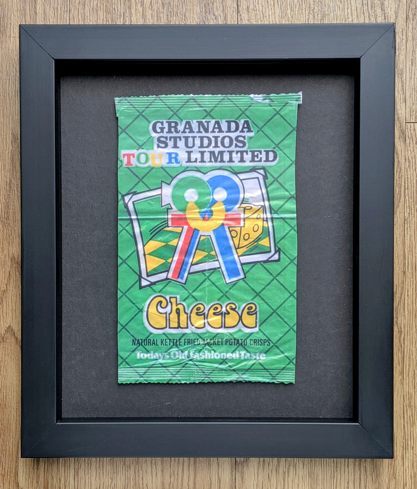 Framed vintage-style green crisps packet with 'Granada Studios Tour Limited Cheese' text on a wooden surface.