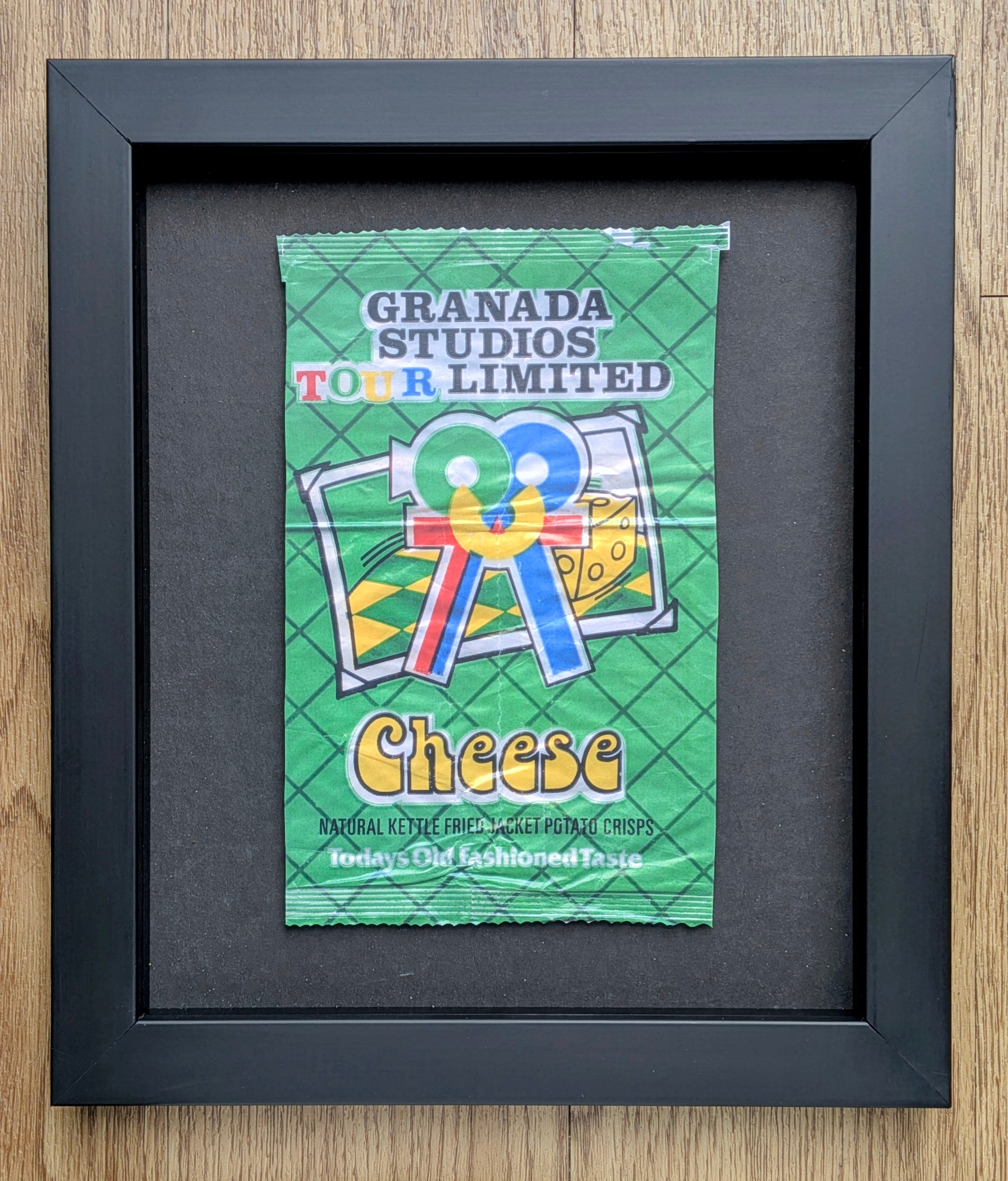 Framed vintage-style green crisps packet with 'Granada Studios Tour Limited Cheese' text on a wooden surface.