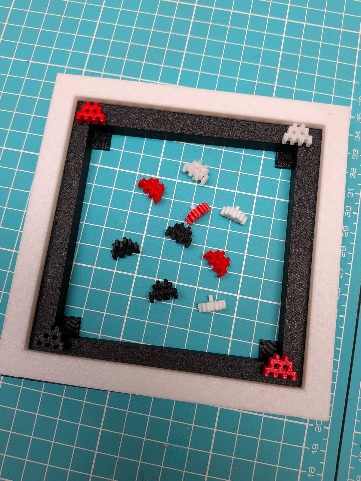 A square 3D-printed frame or tray with a white outer border, black inner border, and small red, black, and white plastic pieces arranged inside on a teal gridded cutting mat.