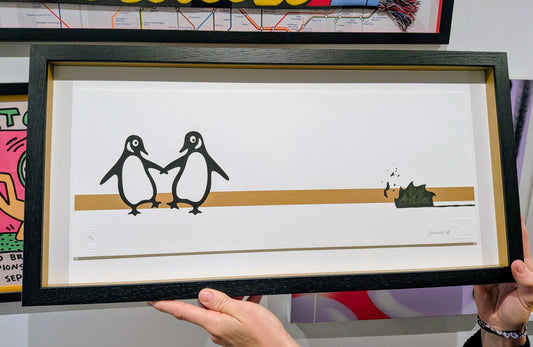 A framed art print featuring two penguins holding hands on a horizontal line, held by hands against a colorful background.