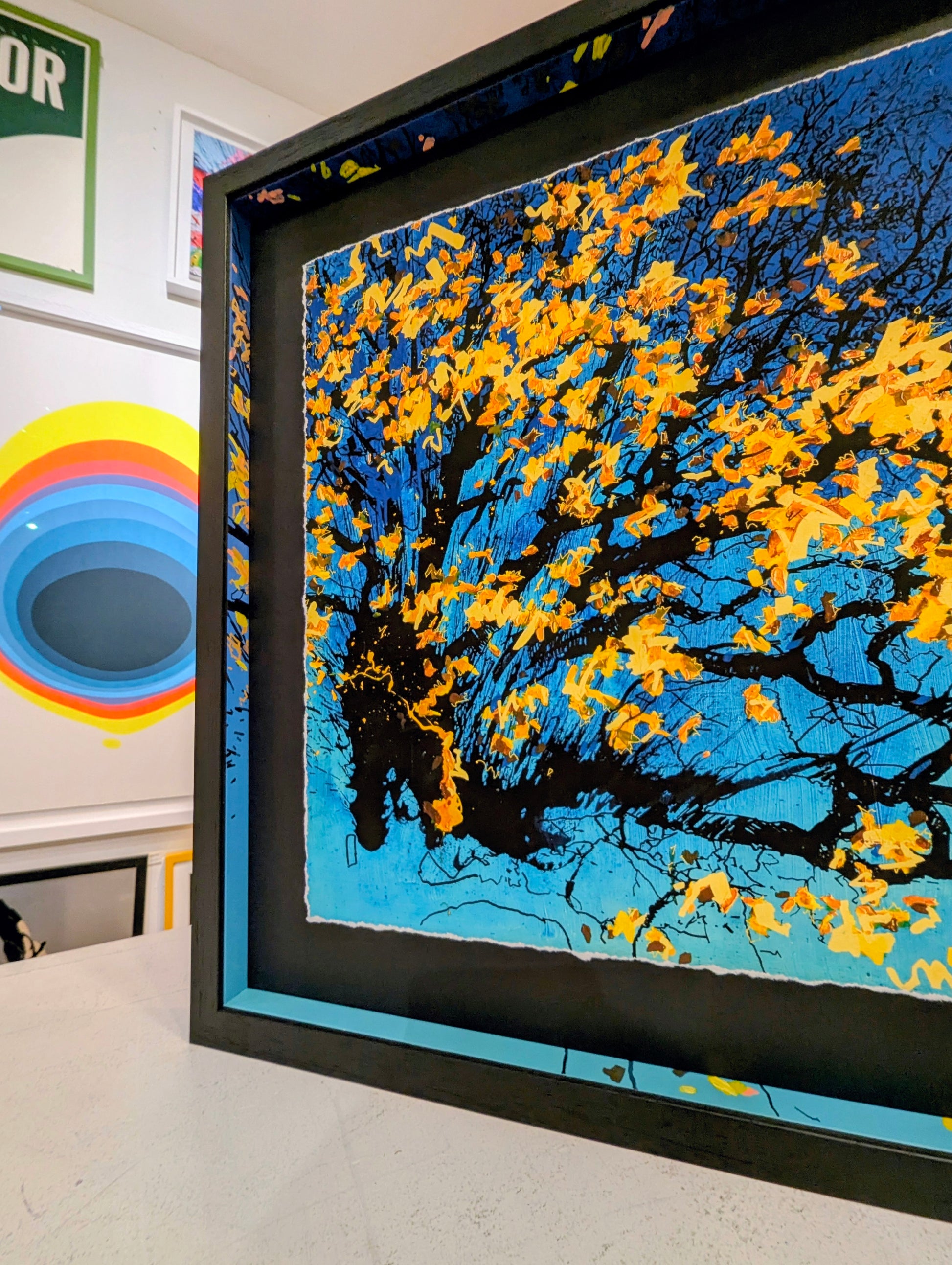 Framed artwork of a tree with yellow leaves against a blue sky, displayed on a table.