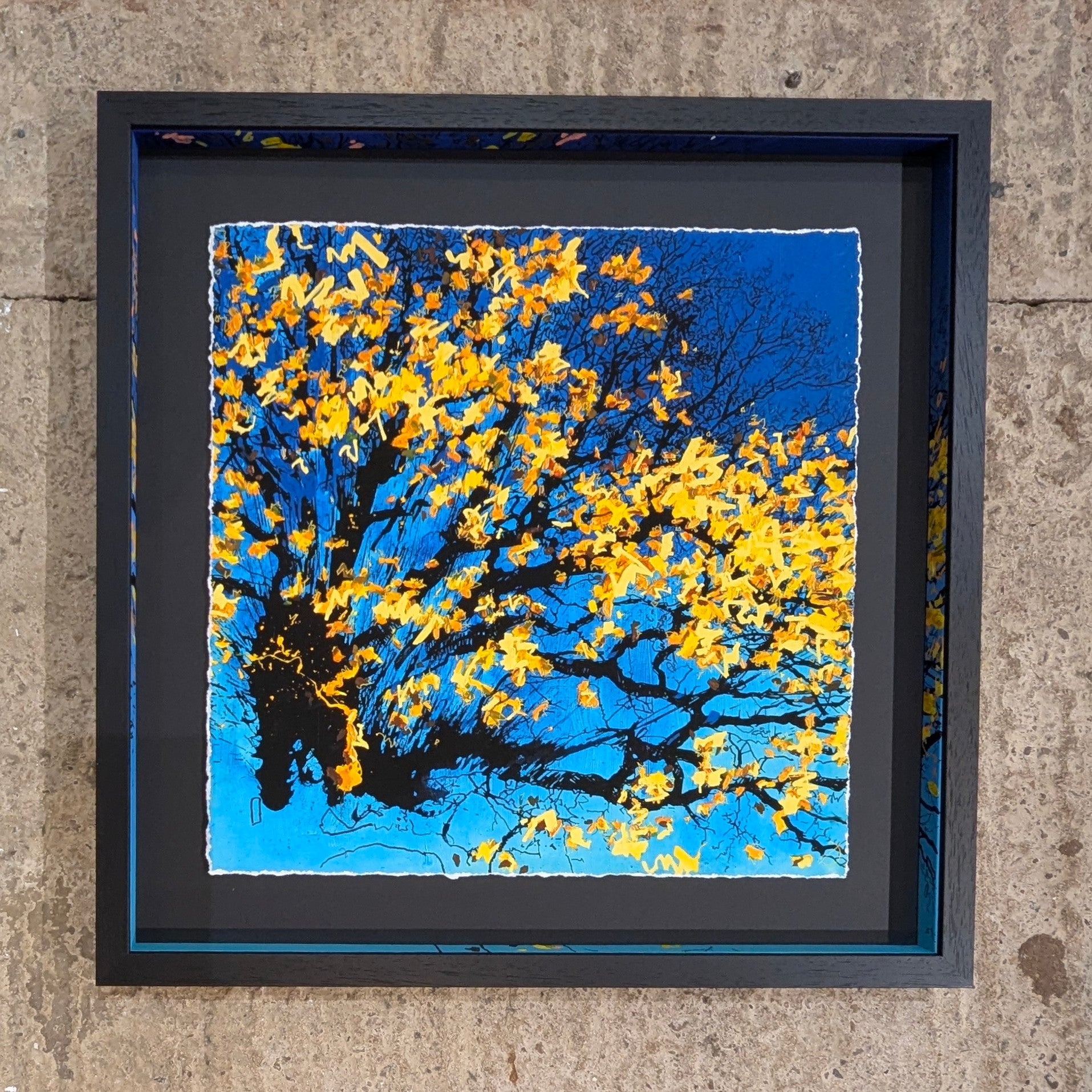 Framed artwork of a tree with yellow leaves against a blue sky on a concrete surface.