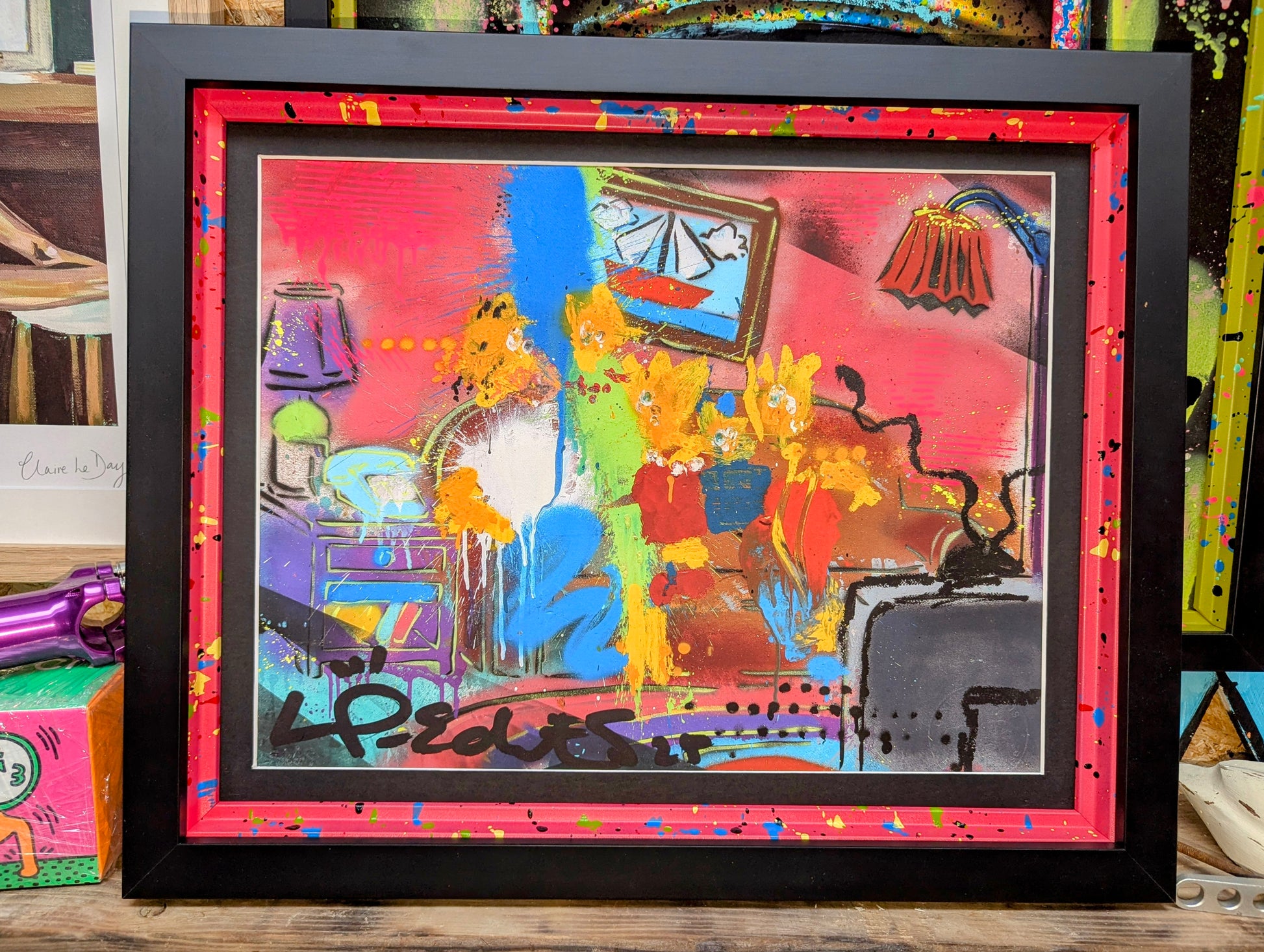 Lp edits artwork framed in a smooth black frame, painted internal frame painted red with blue yellow blue splatters. Artwork is Lp edits chaotic style of The Simpsons, frame is on wooden shelf surrounded by art
