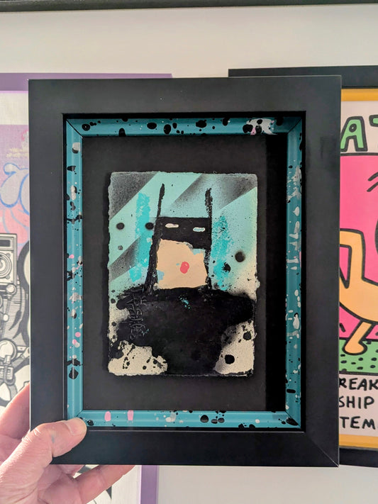 mini cartoon batman in black frame with colourful slips, artwork in background
