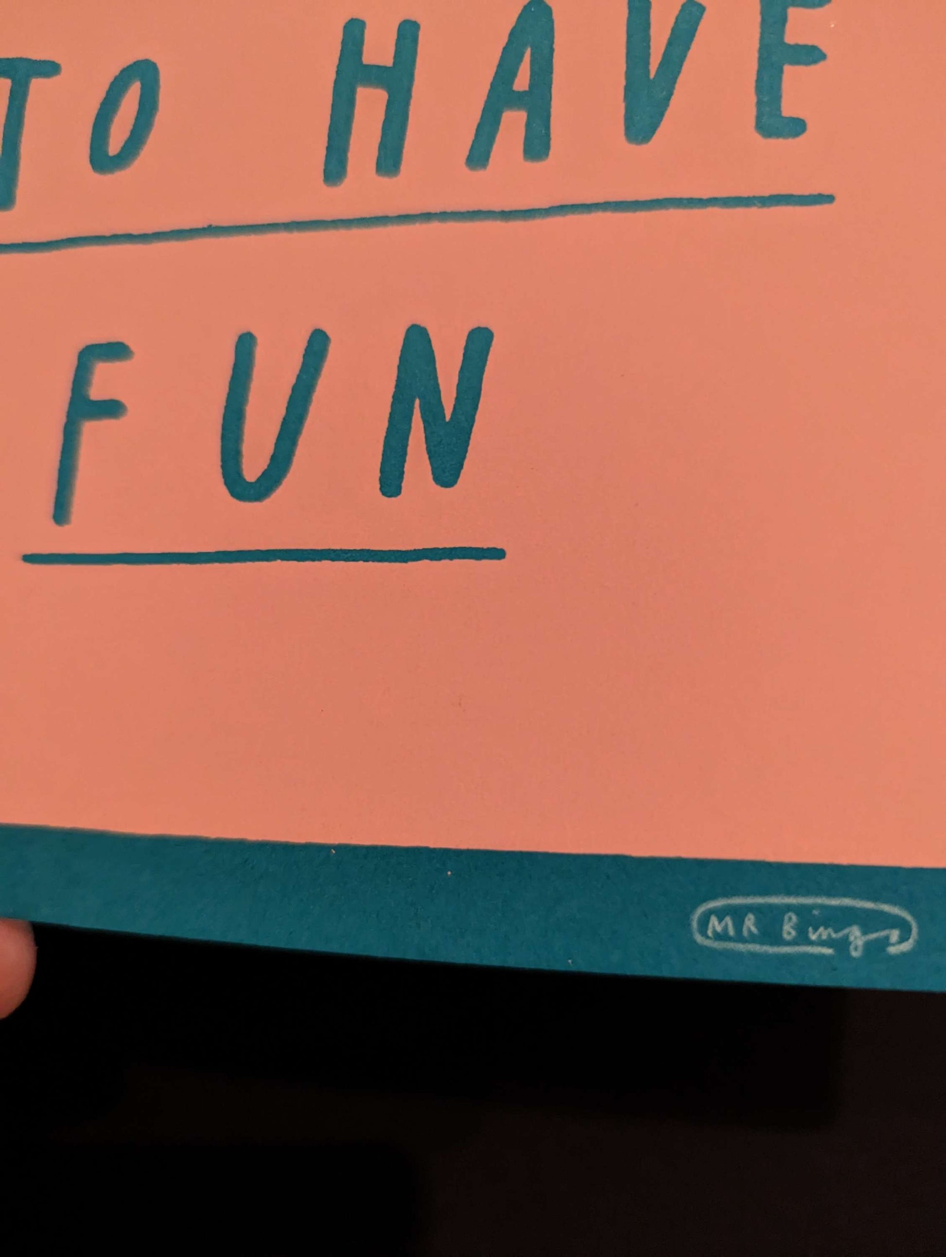 MR BINGO 'DON'T FORGET TO HAVE FUN' Ltd Ed Print, text is hand written style on a salmon pink tombstone against a teal background. card is being held in bottom left corner against a black background, this is a close up of Mr Bingos Signature