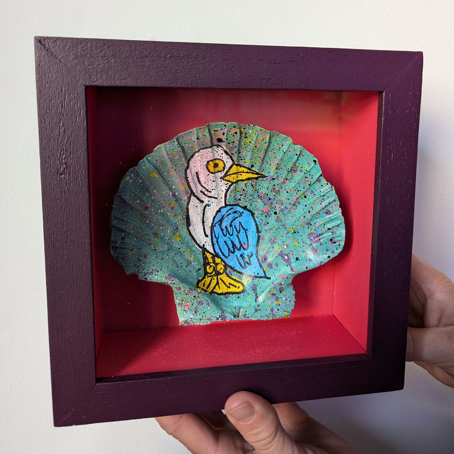 person holding a small purple frame with pink interior. the frame has a sea shell float mounted in the frame, the shell has been splattered with teal and purple and then a cartonn bird none here sid painted on it