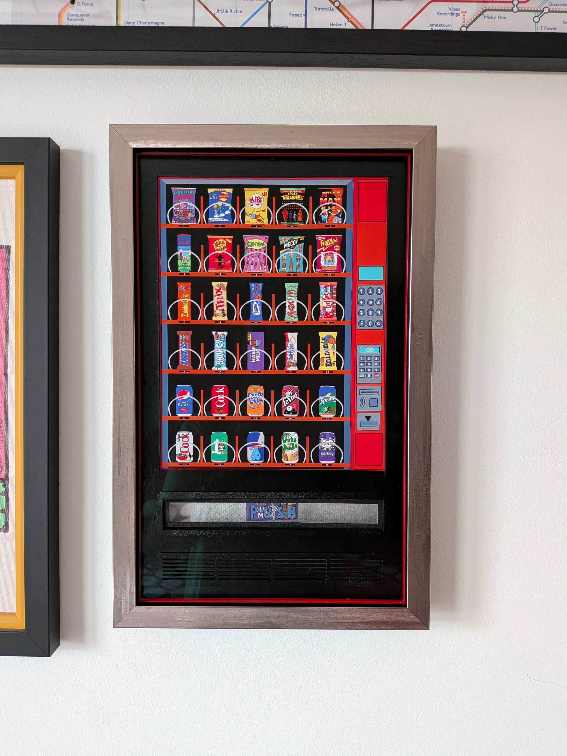 A framed art print depicting a colorful vending machine with various snack packages, mounted on a white wall next to other framed pieces.
