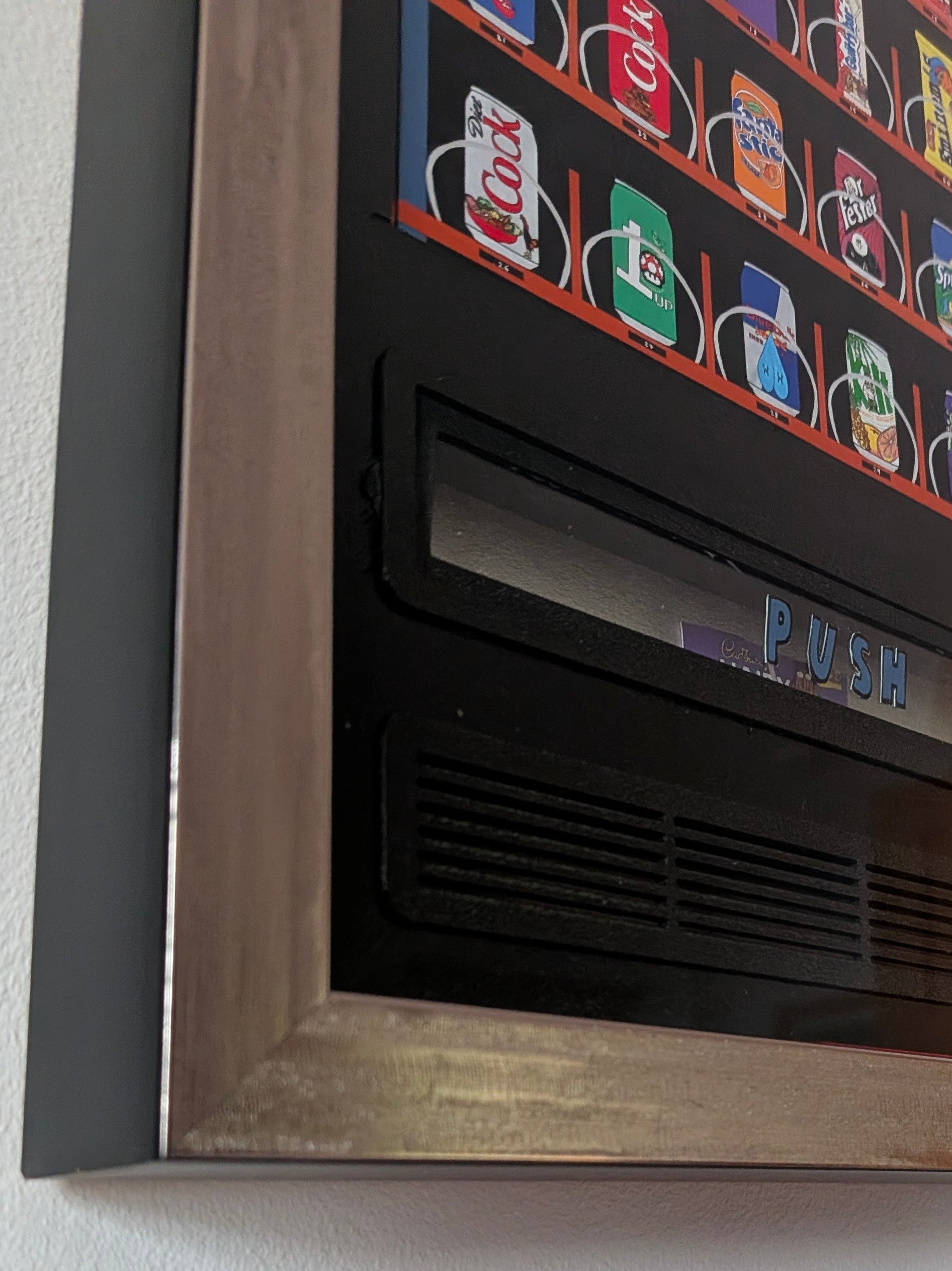 A close-up of a framed wall art print depicting a retro vending machine with colorful beverage cans on a black background, a push slot, and a distressed wood-look frame mounted on a white wall.