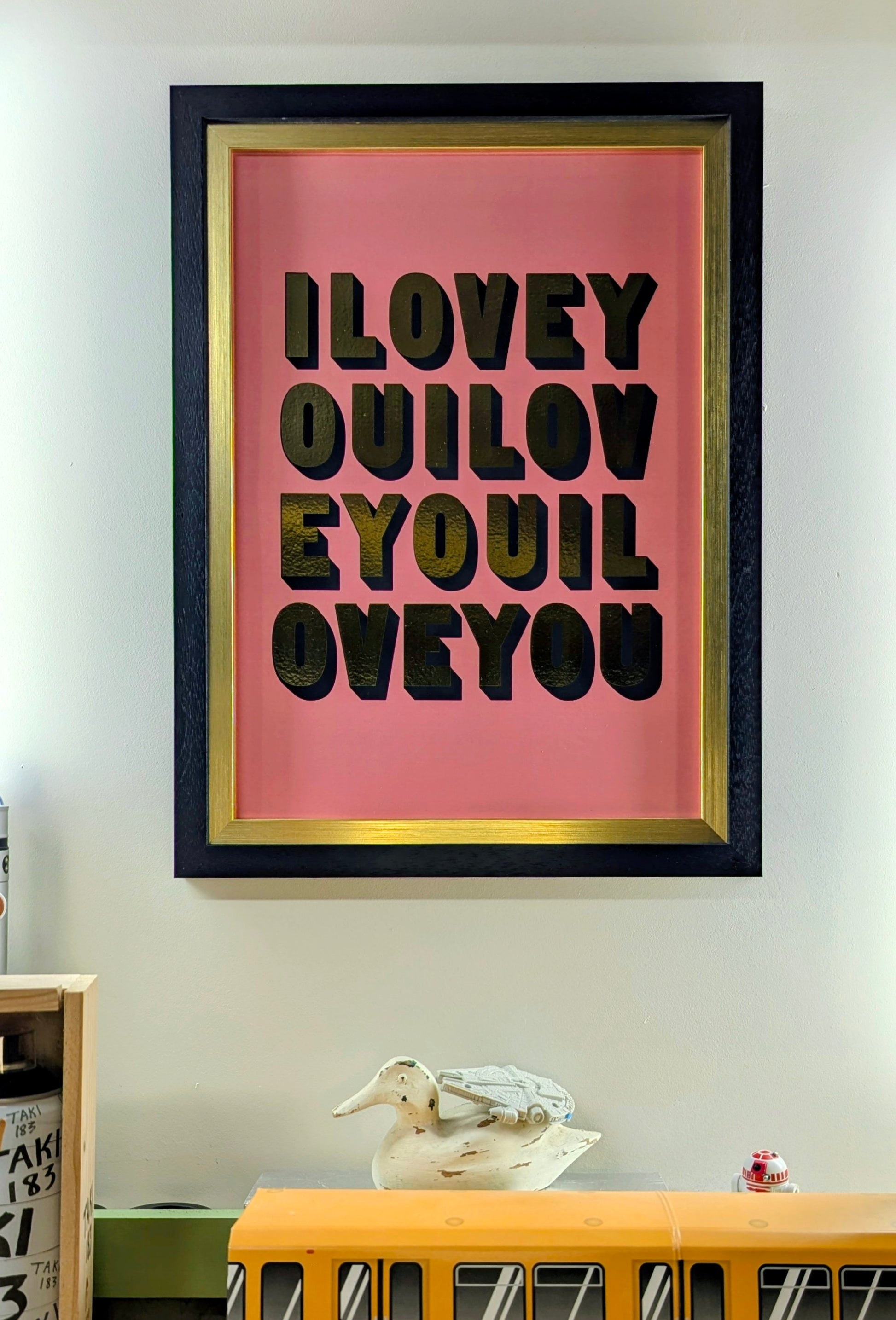 Framed artwork with 'ILOVEYOU' in gold text on a pink background, displayed on a wall. with small items around the base