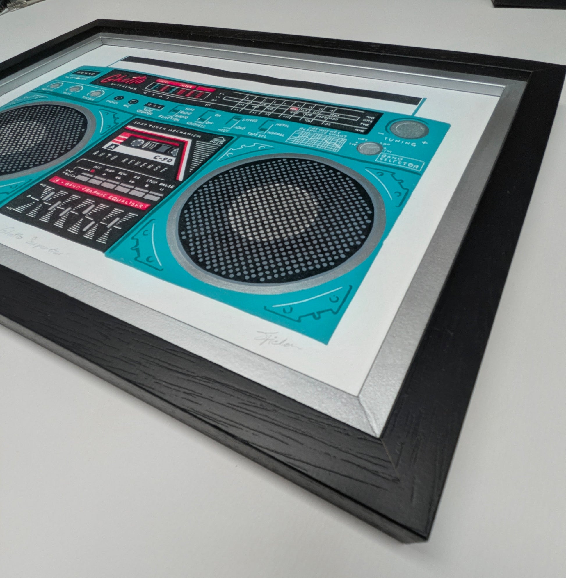 corner of black frame with silver inlay of a retro-style boombox radio artwork  with a turquoise teal body, black speaker grilles