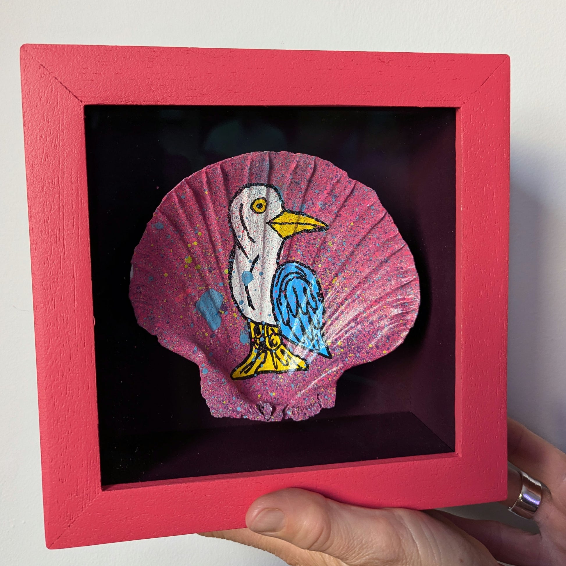 person holding a small pink frame with dark purple interior. the frame has a sea shell float mounted in the frame, the shell has been splattered with teal and purple and then a cartonn bird none here sid painted on it
