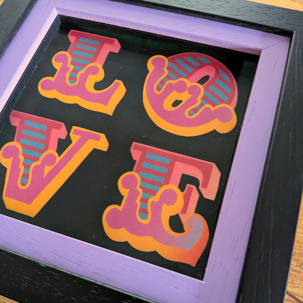 A framed typographic art piece spelling LOVE in stylized letters, with a black background, pink and orange lettering with teal striped accents, a lavender inner frame, and a dark outer frame.