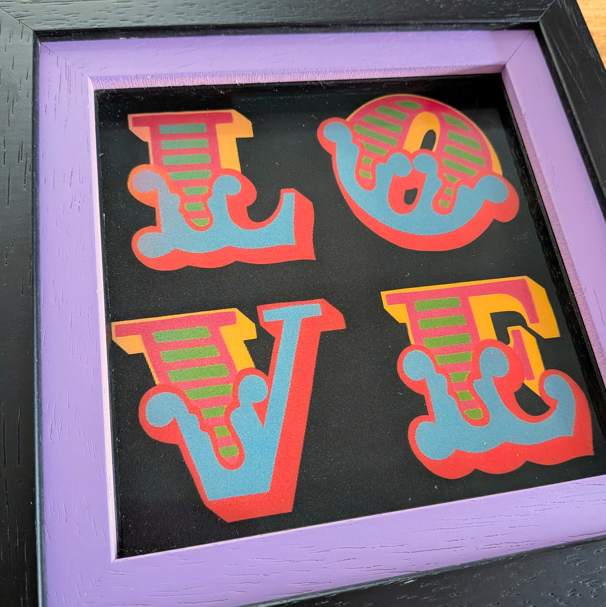 Framed shadow box art featuring the word 'LOVE' in stylized letters. The letters are colored red, light blue, and green with yellow accents, set against a black background and enclosed in a lavender-purple frame.