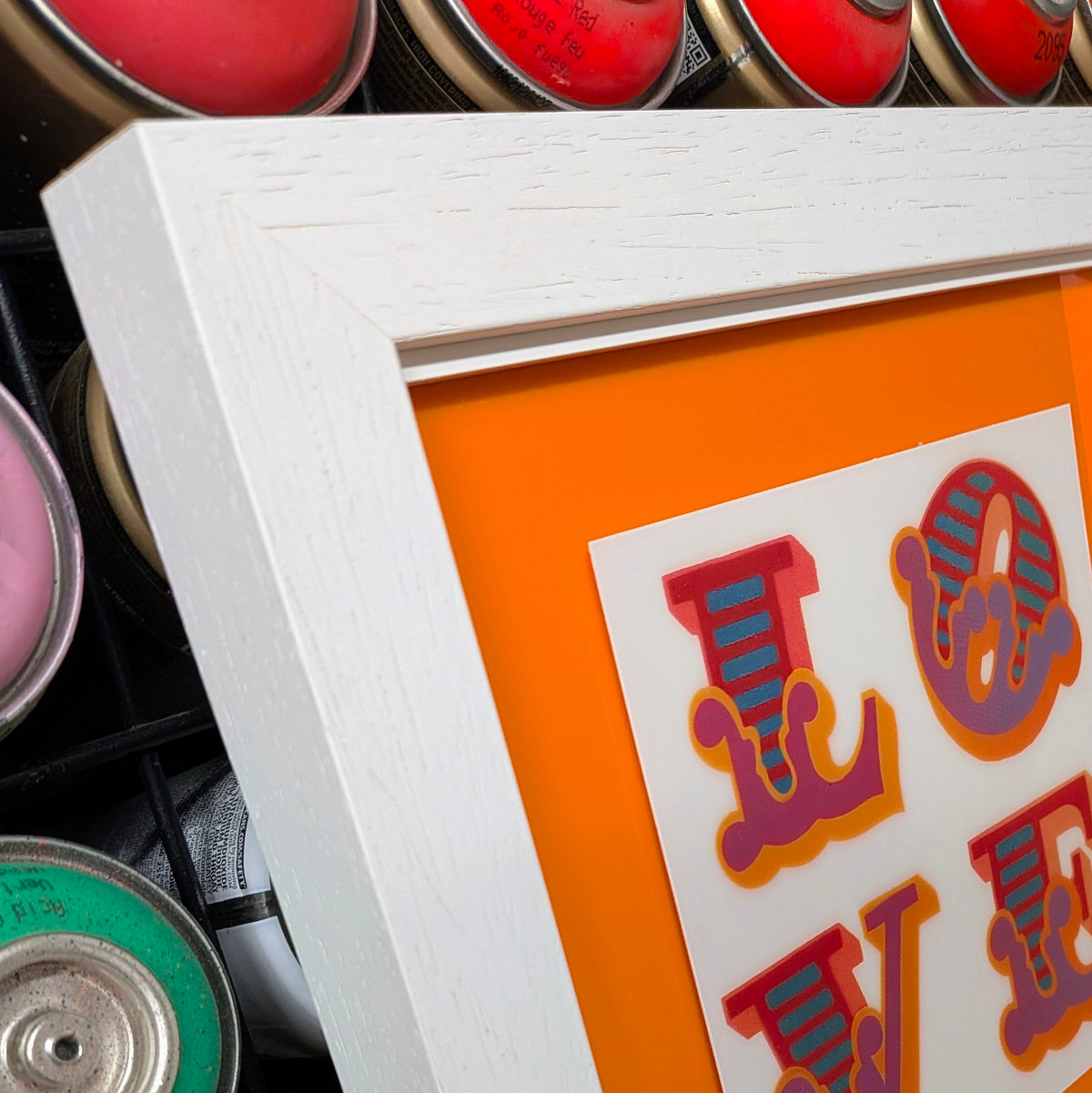 A close-up of a framed art print featuring the word 'LOVE' in stylized red, purple, and blue letters on an orange background, set in a distressed white frame.