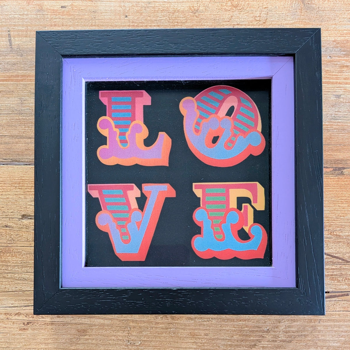 Framed square wall art featuring the word LOVE in stylized retro letters with pink, orange, and blue gradients on a black background, set inside a black frame with a lavender mat on a wooden surface.