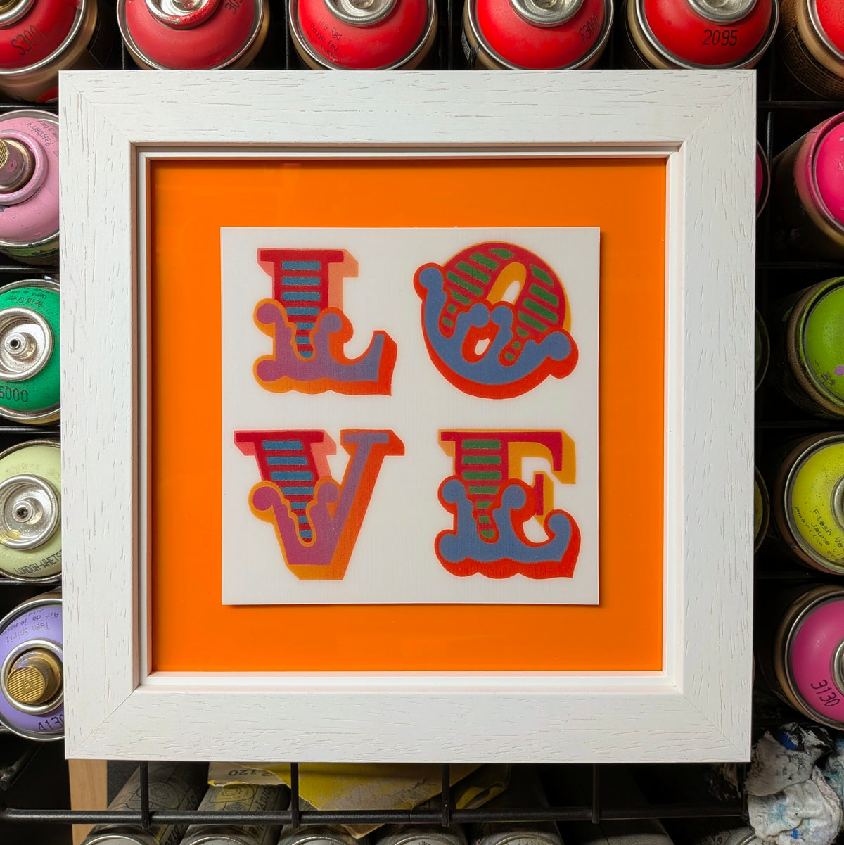 A framed pop-art style 'LOVE' typographic print with retro striped letters in red, orange, blue, and yellow, set against a bright orange background inside a white frame. Spray paint cans are visible in the background.