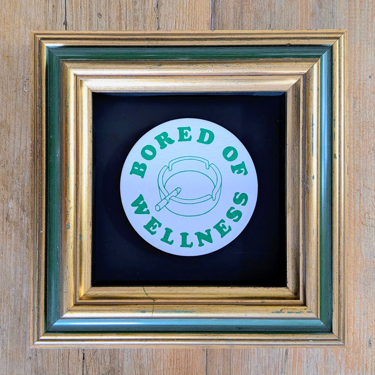 bored of wellness text on beermat on green and gold frame against a wood background.