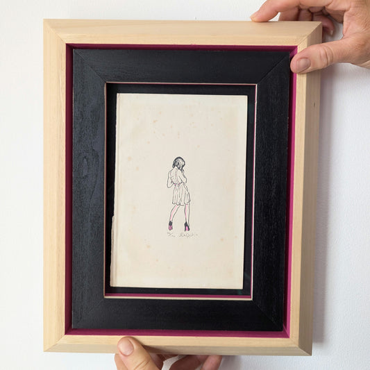 CEAL WARNANTS High Fashion artwork being held, artwork is in a wood frame with black and pink colouring, artwork is drawn on old book paper, the drawing is of a girl wearing a dress, her back is to us and she has pink details to her dress.