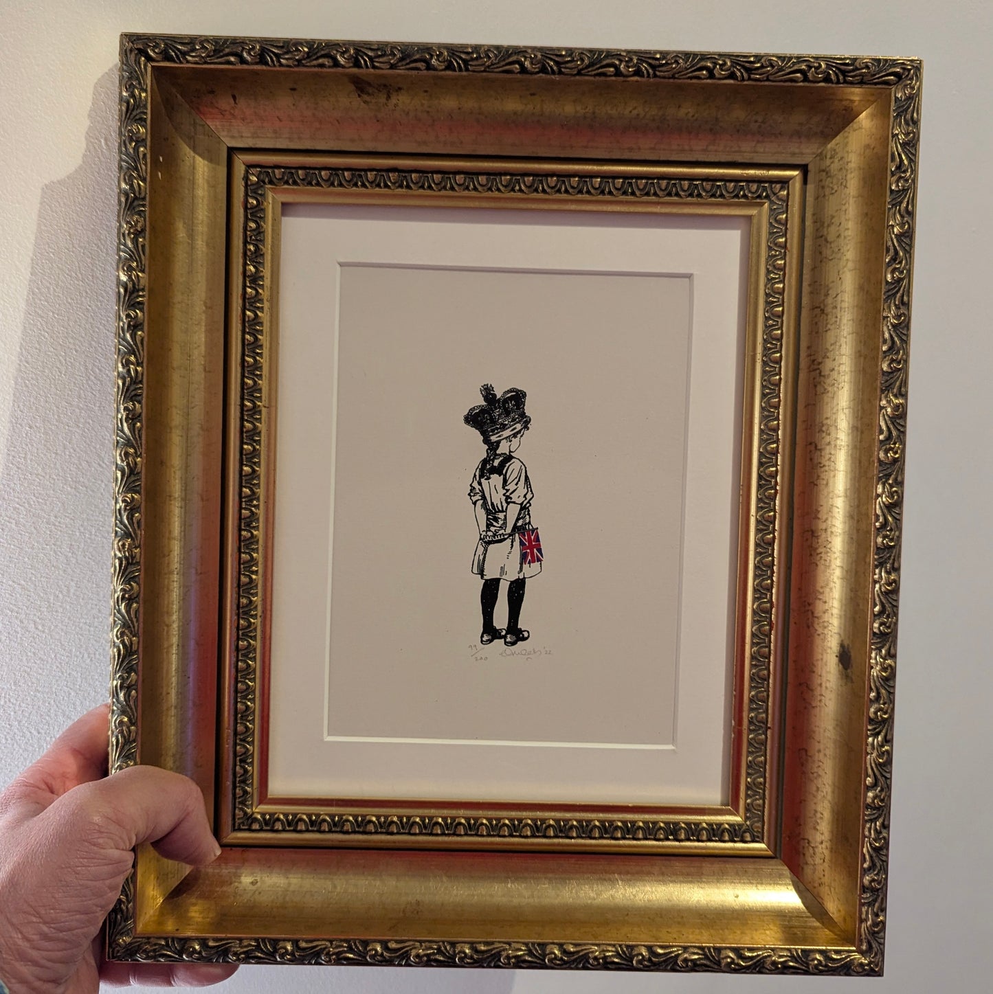 a little girl wearing the royal crown holding a small union jack flag, her back is to us as she look ahead, framed in a gold ornate frame with white mount.