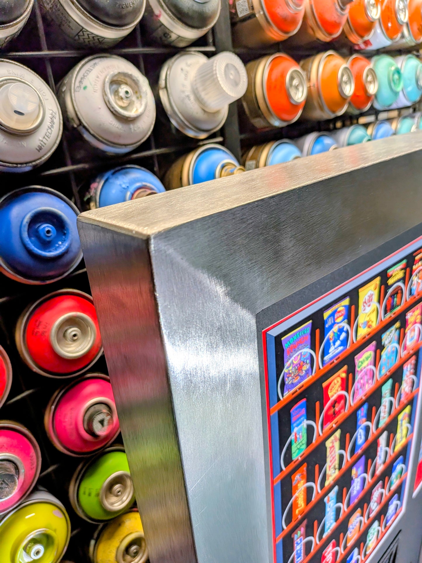 A close-up of a metallic silver vending machine frame and display panel, with colorful spray paint cans visible in the background.