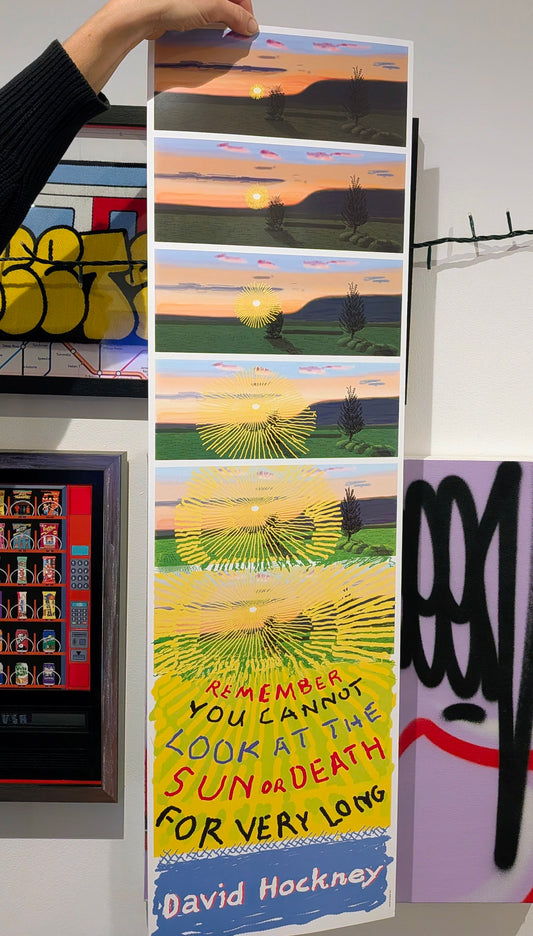 A tall vertical art print featuring a stacked sequence of stylized landscape scenes with a sunset and a bird, with the text 'Remember you cannot look at the sun or death for very long' and 'David Hockney' at the bottom. A hand is holding the top of the print in a gallery-like setting with other graffiti-style artworks visible.