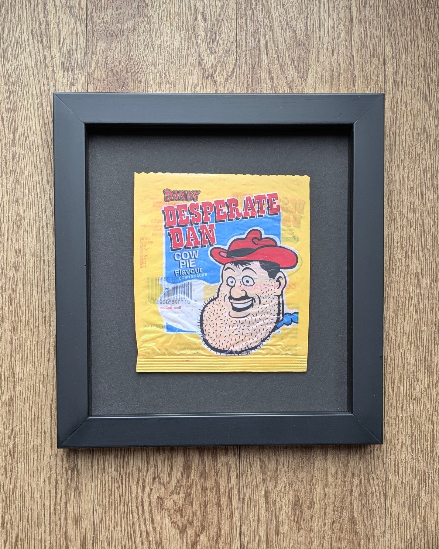 Framed Desperate Dan candy wrapper on a wooden surface