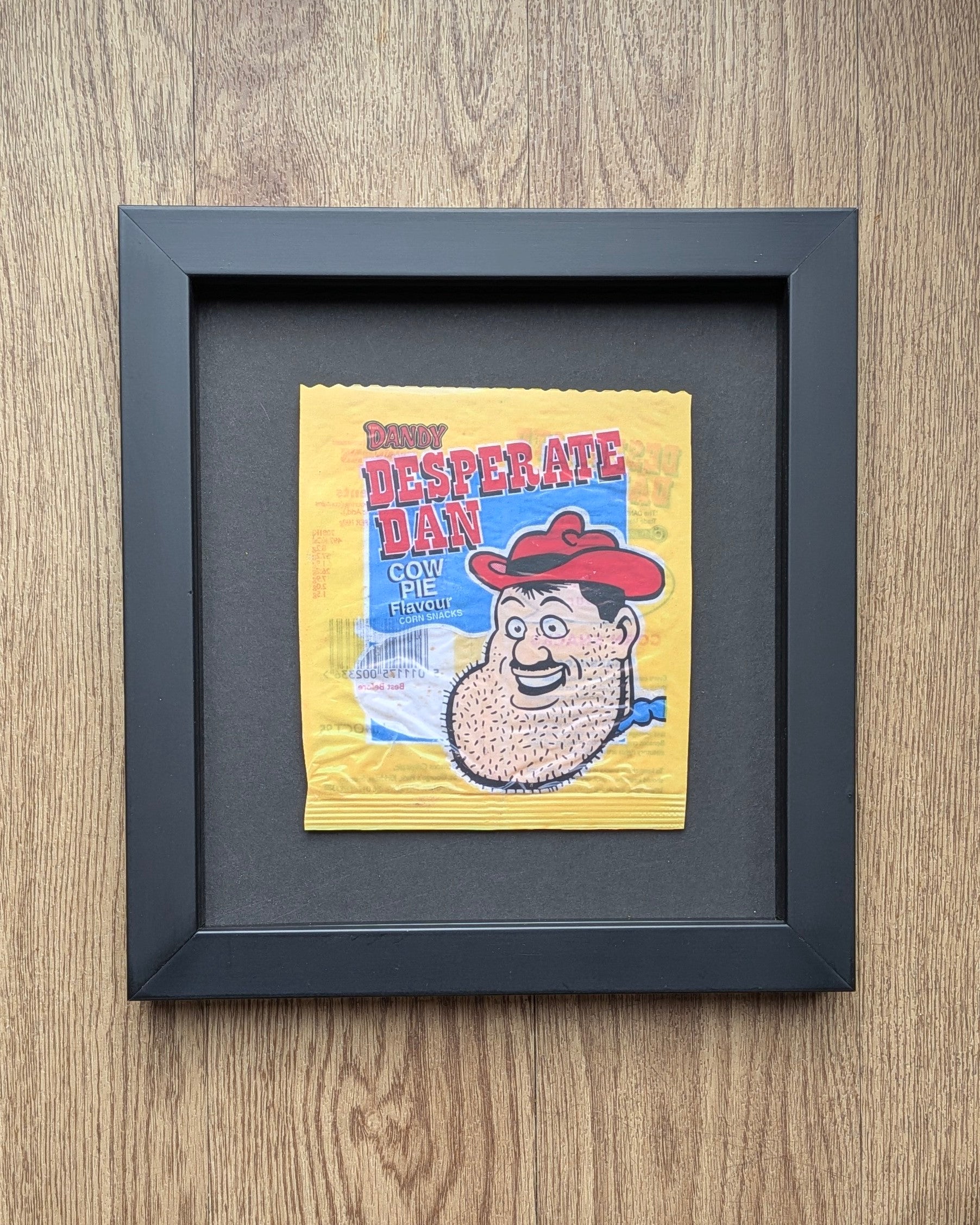 Framed Desperate Dan candy wrapper on a wooden surface