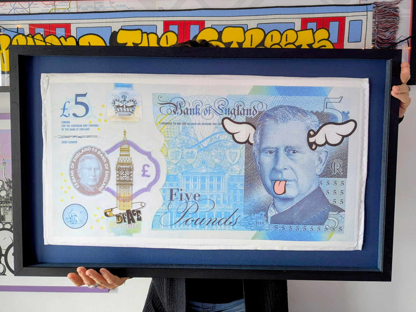 Framed artwork of a five-pound note with dface cartoonish elements, artwork in background