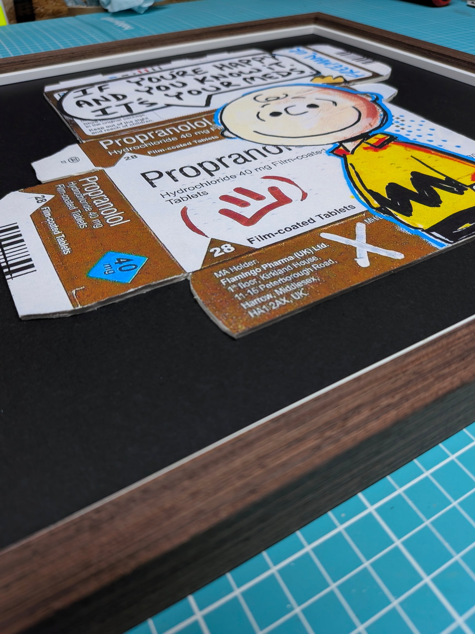 A framed collage artwork featuring a black mat, dark wood frame, and multiple printed inserts including a Propranolol card, a handwritten note, and a cartoon illustration, photographed on a teal cutting mat.