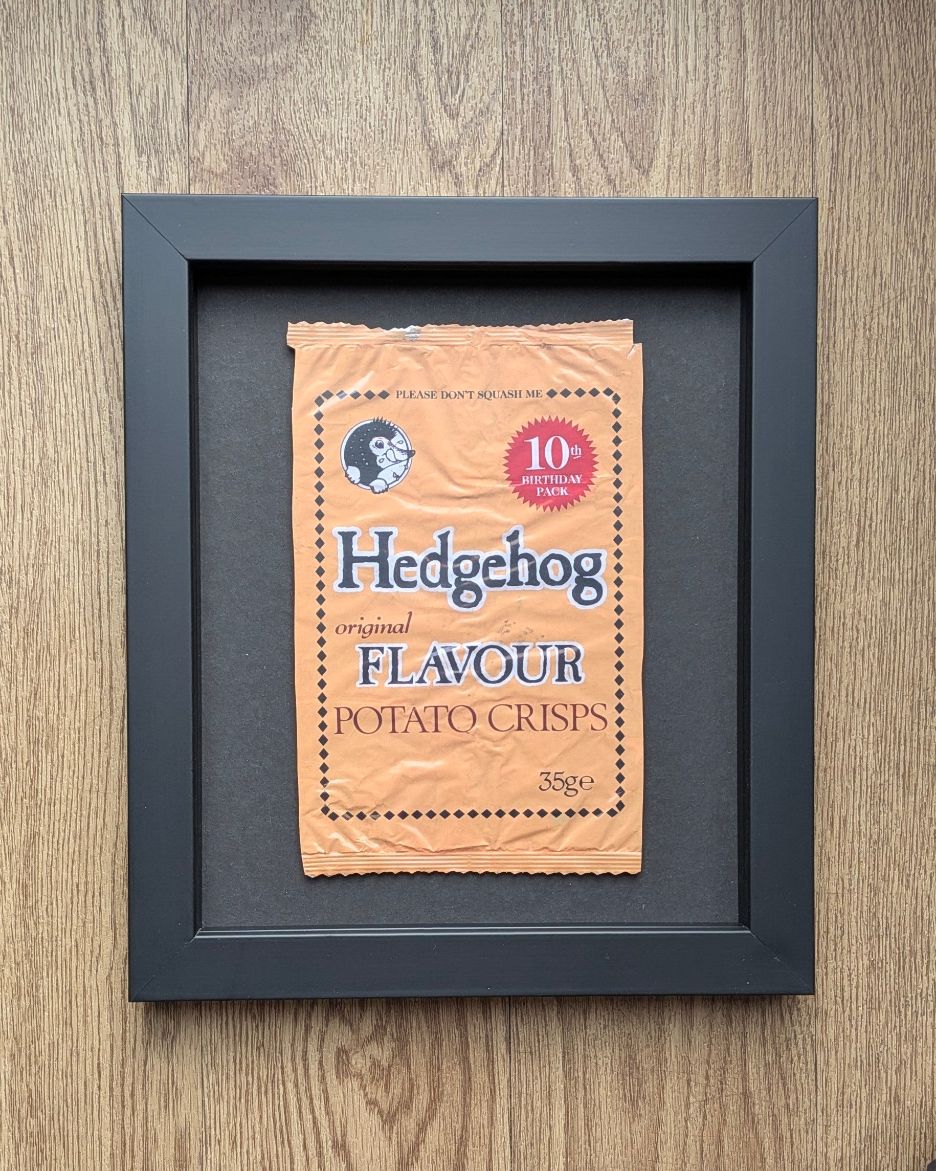 Framed picture of a vintage-style 'Hedgehog Flavour' potato crisps packet on a wooden surface.