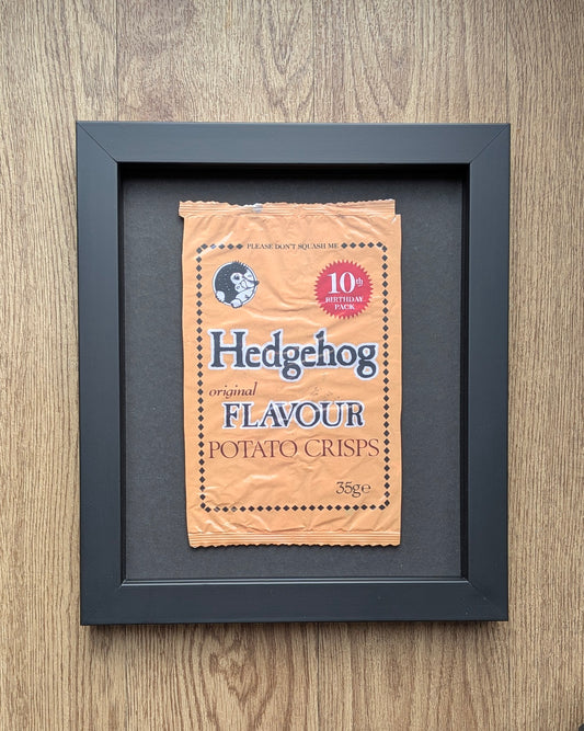 Framed picture of a vintage-style 'Hedgehog Flavour' potato crisps packet on a wooden surface.