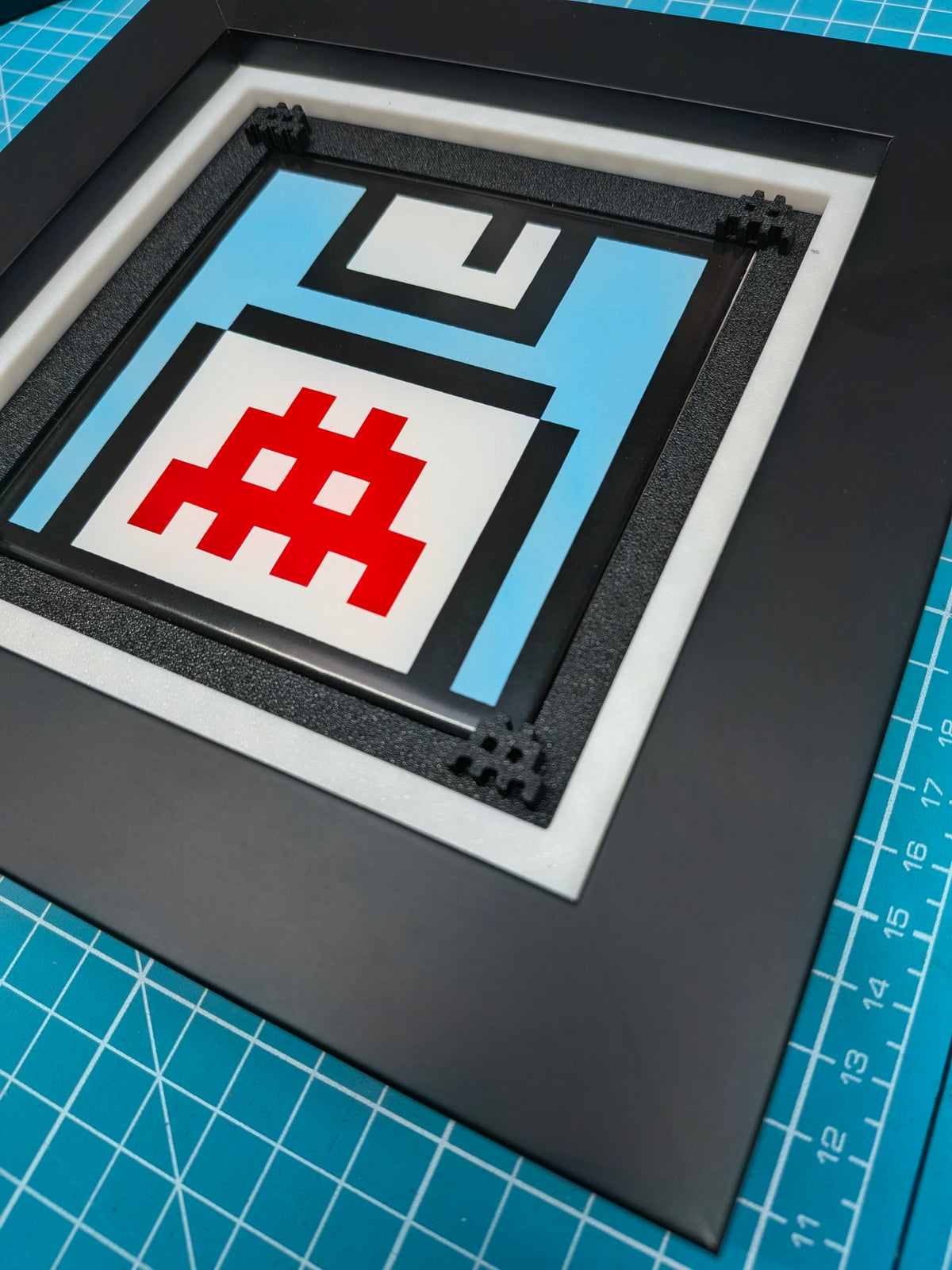 A square black-framed art print or decorative plaque featuring a stylized retro floppy disk design in light blue, white, black, and red, photographed on a blue gridded cutting mat.