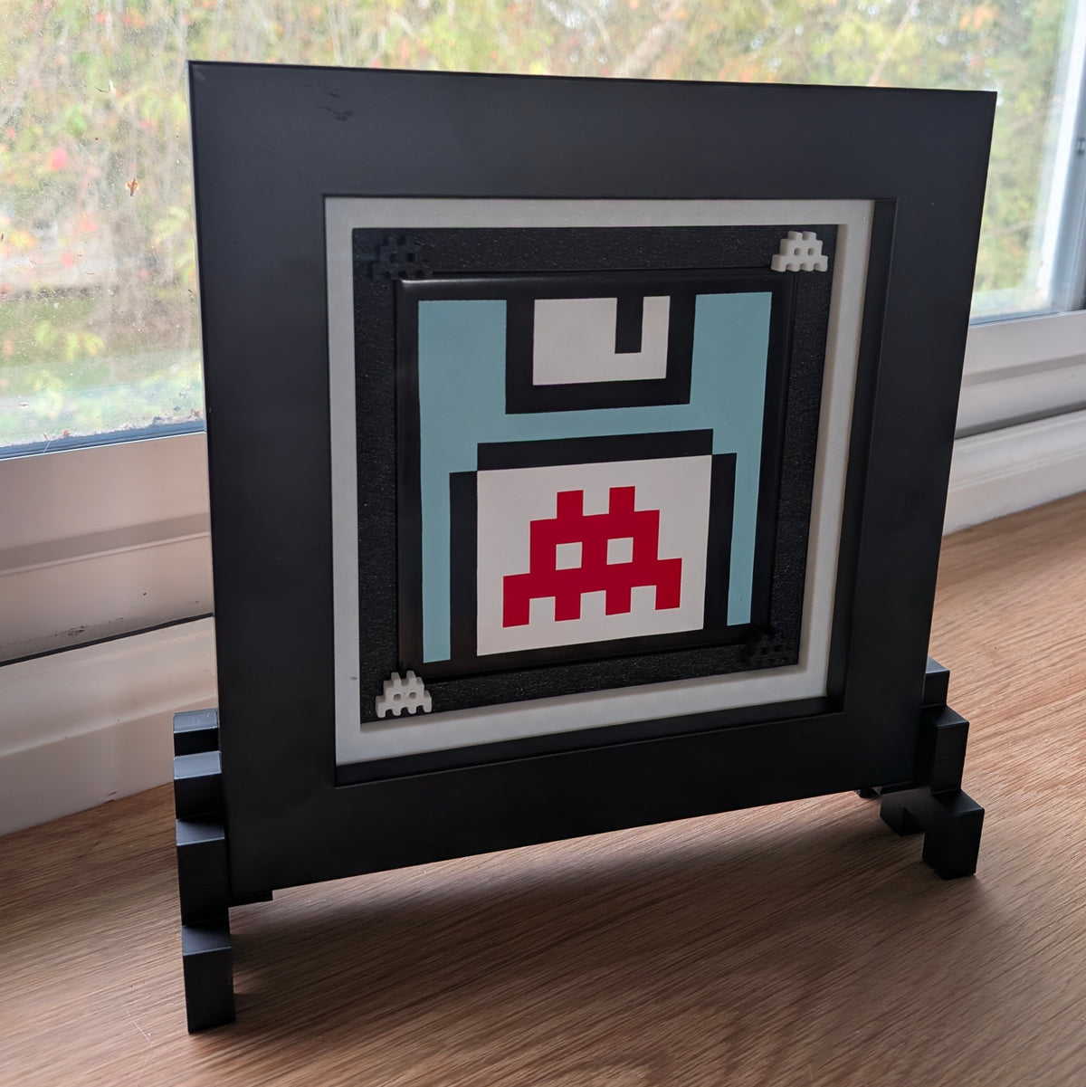 A square black picture frame with a 3D block-style stand, holding a pixel-art floppy disk design in light blue, white, black, and red, displayed on a wooden floor near a window.