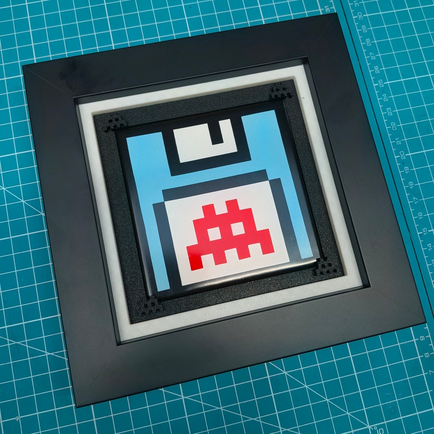 A square black shadowbox frame containing a pixel-art floppy disk design in light blue, white, and red, displayed on a green cutting mat.